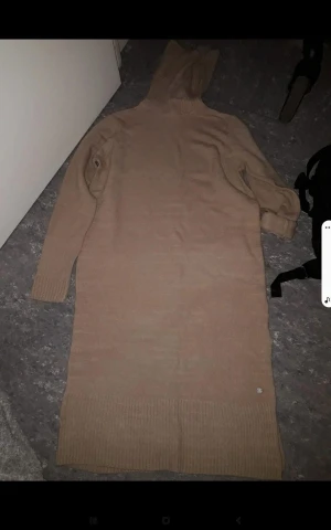 Beige stickad långklänning med luva - Mysig beige stickad långklänning med luva och långa ärmar. Klänningen har ribbade muddar vid ärmslut och nederkant, perfekt för en chill och avslappnad look. Enkel och stilren design som är lätt att matcha med sneakers eller boots.