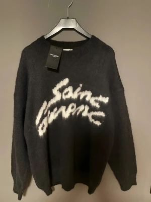 Saint Laurent Paris Mohair Logga Sweater – Svart (Unisex) - Jag svarar gärna på alla frågor du kan ha, och priset är förhandlingsbart. Tveka inte att höra av dig! Gör dig redo att addera en touch av lyx till din stil med denna fantastiska tröja.  Oversize svart mohairblandad tröja med en tydlig vit, fluffig Saint Laurent-logga över bröstet. Mjuk, varm och lätt lurvig textur som är typisk för mohairstickningar. Ribbstickad halsringning, muddar och fåll.