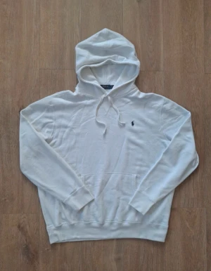 Vit hoodie från Polo Ralph Lauren - Vit hoodie från Polo Ralph Lauren med klassisk blå logobrodyr på bröstet. Tröjan har huva med snörning, magficka och långa ärmar. Materialet är mjukt och består av 60% bomull och 40% polyester, perfekt för chill dagar.