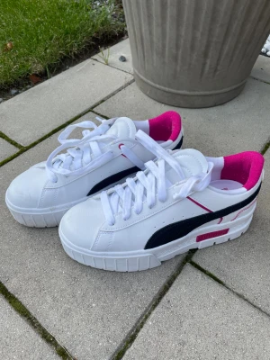Vita Puma sneakers med rosa detaljer - Snygga vita sneakers från Puma med svart sidodetalj och rosa insida samt rosa logga på sulan. Skorna har rund tå, platt sula och klassisk snörning. Perfekta för dig som gillar sportig stil med färgklick. Materialet är skinn och syntet.