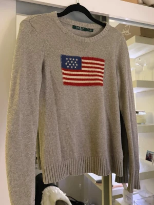 Grå stickad tröja Lauren Ralph Lauren - Snygg grå stickad tröja från Lauren Ralph Lauren med amerikansk flagga broderad på bröstet. Tröjan har rund hals, ribbade muddar och långärmad passform. Perfekt för dig som gillar klassisk stil med en twist. Säljer min syrras tröja.