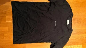 Svart t-shirt från Les Deux - Svart t-shirt från Les Deux med enkel vit logga tryckt på bröstet. Klassisk rund halsringning och korta ärmar. Tillverkad i mjuk bomull för en skön känsla. Perfekt basic-plagg med stilren look.