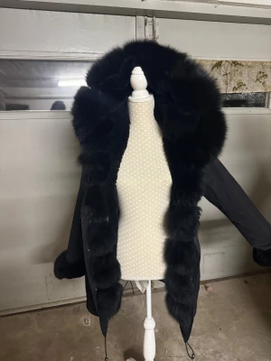 Svart parkajacka med pälsdetaljer - Snygg svart parkajacka med fluffig päls runt huvan, ärmsluten och längs framkanten. Jackan har långa ärmar, dragsko i midjan och huva. Materialet är slitstarkt och pälsen ger en lyxig vibe. Perfekt för kalla dagar när du vill vara både varm och stilren. Storlek XS men passar även S.