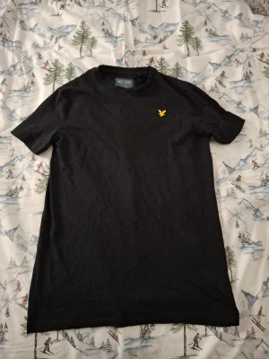 Svart t-shirt från Lyle & Scott - Svart t-shirt från Lyle & Scott med klassisk rund hals och korta ärmar. På bröstet sitter den gula fågeln som broderad detalj. T-shirten är gjord i mjuk bomull och har en clean, stilren look som funkar till allt.