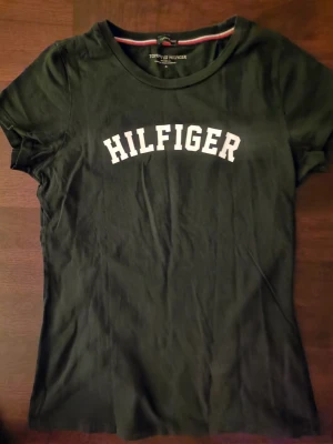 Svart t-shirt från Tommy Hilfiger - Svart t-shirt från Tommy Hilfiger med vit HILFIGER-text tryckt över bröstet. Klassisk rund halsringning och korta ärmar 💕