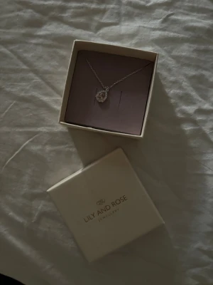Silver halsband från Lily and Rose - Snyggt halsband i silver från Lily and Rose med en droppformad berlock. Berlocken har en ljusrosa sten i mitten och är omgiven av glittriga små stenar. Perfekt för dig som gillar eleganta och stilrena smycken med lite extra bling.