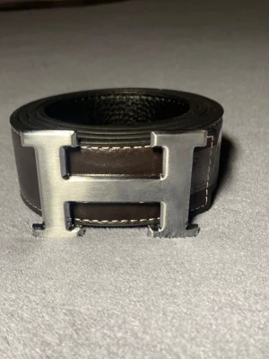 Svart skinnbälte med H-spänne - Säljer ett svart bälte med ett stort H-format metallspänne i silverfärg. Bältet är gjort av äkta skinn och har vita sömmar längs kanterna som ger en snygg kontrast. Perfekt accessoar för att lyfta din outfit.