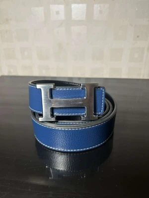 Blå skinnbälte med H-spänne - Säljer ett stilrent blått bälte med ett stort H-format metallspänne. Bältet är gjort i skinn med vit kontrastsöm längs kanterna och har en klassisk, modern look som passar till jeans eller kostym. Perfekt accessoar för att lyfta din outfit.