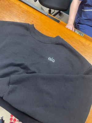 Svart sweatshirt från Alo, storlek S - Svart sweatshirt från Alo med diskret logga broderad på bröstet. Klassisk rund halsringning och ribbade muddar vid ärmslut och nederkant. Tillverkad i en mjuk bomulls- och polyestermix som känns skön mot huden.