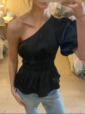 Svart oneshoulder topp med puffärm - Säljer en svart oneshoulder topp med en voluminös puffärm och markerad midja med resår. Toppen har en lätt glansig finish och peplum-liknande nederdel som ger en snygg siluett. Perfekt för dig som vill sticka ut lite extra.