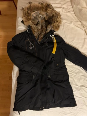 Svart parajumper med päls och detaljer - Säljer en svart parajumper jacka i storlek S med päls, modellen går över rumpan. Pälsen är skadad på få ställen på insidan av luvan (bild) samt ett hål i fickan. Annars är jackan i mycket bra skick. 