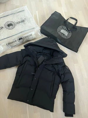 Svart Wyndham Parka från Canada Goose - Svart Wyndham Parka från Canada Goose med huva och ikonisk patch på ärmen. Jackan är dunfodrad, har långa ärmar, dragkedja och knappar framtill. Perfekt för kalla dagar och har en clean, stilren look som funkar till streetwear. Snyggt quiltad design och riktigt varm.