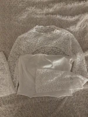 Vit spetsblus med lång ärm - Säljer en vit blus i transparent spets med långärmade armar och hög hals. Blusen har ett vackert blommigt spetsmönster och är croppad i modellen. Perfekt för dig som gillar romantiska detaljer och vill ha en feminin look.