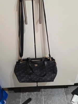 Svart handväska från Lycke Oslo - Snygg svart handväska från Lycke Oslo med logomönster över hela väskan och guldfärgade detaljer. Väskan har både handtag och justerbar axelrem samt dragkedja upptill. Perfekt storlek för allt du behöver ha med dig och riktigt stilren look.