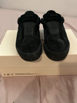 Svarta sneakers Axel Arigato mocka - Säljer ett par svarta sneakers från Axel Arigato i storlek 39. Skorna är tillverkade i mjuk mocka med rund tå och platt sula. Stilren design utan snörning, med logga på plösen. Kommer med originalkartong och dustbag.