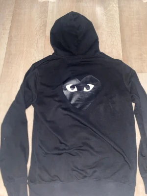Cdg - Svart CDG hoodie i bra skick. Klassisk modell från Comme des Garçons med stilren design. Hoodien är väl omhändertagen, sparsamt använd och utan fläckar eller hål. Mjuk och bekväm passform, perfekt för både vardag och streetwear-look.