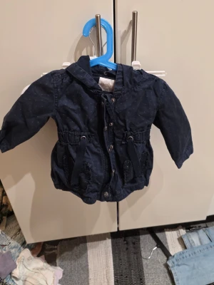 Mörkblå prickig parkajacka Baby by Lindex - Säljer en mörkblå parkajacka från Baby by Lindex i storlek 86. Jackan har små ljusblå prickar, huva, knappar och snörning i midjan. Materialet är bomull och jackan har långa ärmar. Perfekt för dig som vill ha en söt och praktisk jacka till de små.