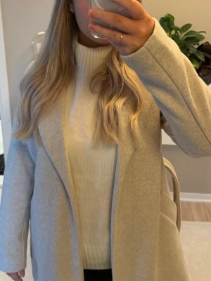 Beige kappa med bälte från Vero Moda - Snygg beige kappa från Vero Moda med bälte i midjan. Jackan har två fickor framtill och är långärmad med en stilren, rak passform. Använd fåtal ggr💕passar xs och s 