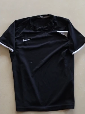 Svart Nike tränings tshirt L - Svart träningsväst från Nike med vit logga och vita detaljer vid ärmslut och axlar. Västen är gjord i ett lätt och funktionellt syntetmaterial som andas, perfekt för sport och aktivitet. Enkel och stilren design med rund hals.