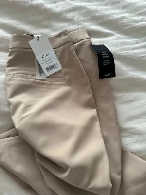Beige kostymbyxor från NA-KD - Snygga beige kostymbyxor från NA-KD med rak passform och slits vid benslutet. Byxorna har hög midja och är tillverkade i ett mjukt material som ger en clean look. Perfekta att styla med en oversized skjorta eller kavaj. Aldrig använd 