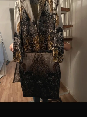 Apanage Collection Matching Blazer & Skirt Set – Baroque Pattern - Elegant two-piece set from Apanage Collection, consisting of a matching blazer and skirt. The set features a striking baroque-inspired pattern in black, gold, beige, and cream tones. Classic, tailored design that looks sophisticated and timeless. Pieces can be worn together for a polished look or styled separately. High-quality fabric with a slight stretch for comfort. Perfect for formal events, work, dinners, or special occasions. Details: Brand: Apanage Collection Item: Blazer + Skirt set Size