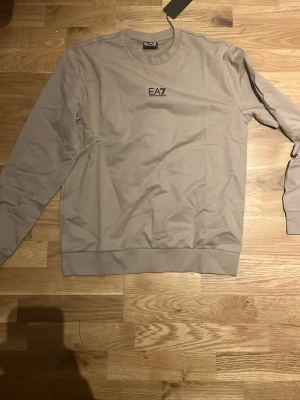 Beige sweatshirt från EA7 Emporio Armani - Snygg beige sweatshirt från EA7 Emporio Armani med diskret logga på bröstet och svarta ränder i nacken. Tröjan har rund hals, långa ärmar och är tillverkad i mjuk bomull. Perfekt för en clean och stilren look.