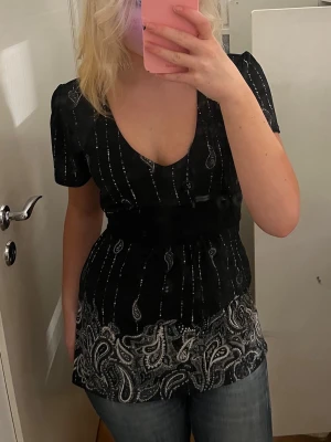 Svart blus med paisleymönster - Snygg svart blus med djup v-ringning och korta puffärmar. Blusen har ett vitt paisleymönster nertill och tunna glittriga ränder längs hela plagget. Figurnära passform som markerar midjan och ger en feminin siluett.