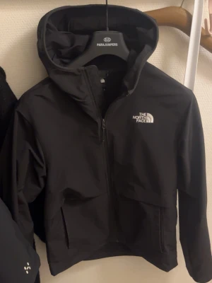 The North Face Vindjacka - Svart vindjacka från The North Face med huva och dragkedja framtill. Jackan har en stilren design med logga på bröstet och ryggen. Tillverkad i polyester, perfekt för blåsiga dagar. Passar dig som gillar funktion och enkel stil. En liten ljusblå färgfläck på insidan som inte märks av när du använder den så inga mera skador eller problem än det! 🔥 tveka inte på att ställa en fråga eller två vid intresse!!❤️👍