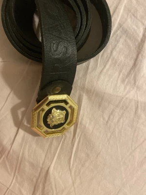 Svart bälte med guldspänne Versace - Säljer ett svart bälte från Versace i skinn med ett stort, oktagonformat guldspänne och ikoniskt Medusa-huvud. Bältet har präglad logga längs remmen och känns riktigt lyxigt och statement.