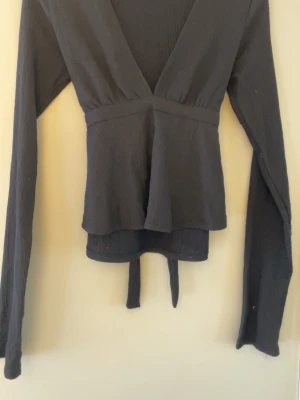 Svart omlottopp från Nelly - (Dåligt ljus på bilden) Säljer en svart omlottopp från Nelly i storlek M. Toppen har lång ärm, v-ringad hals och knytband i midjan som ger en snygg siluett. Materialet är lätt strukturerat och känns mjukt mot huden. Perfekt att styla med jeans eller kjol.