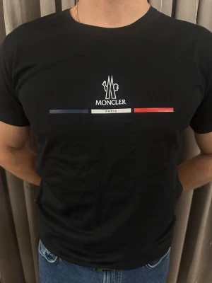 Svart Moncler Paris t-shirt - Säljer en svart t-shirt från Moncler med tryck i vitt, blått och rött på bröstet. T-shirten har rund hals och korta ärmar. Materialet är mjuk bomull och passformen är normal. Perfekt för dig som gillar stilrena designer och exklusiva märken.