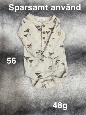 Body med svampmönster från Newbie - Söt body från Newbie i ljus beige med tryck av olika svampar i brunt, gult och grått. Bodyn har långa ärmar, träknappar framtill och praktiska tryckknappar i grenen. Perfekt för små äventyrare!🌸Sparsamt använd. Storlek 56