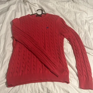 Röd kabelstickad tröja från Morris - Säljer en snygg röd kabelstickad tröja från Morris i storlek M. Tröjan har rund hals, långa ärmar och ribbade muddar vid ärmslut och nederkant. Perfekt för dig som gillar klassisk stil med en modern twist. Diskret Morris-logga broderad på bröstet.
