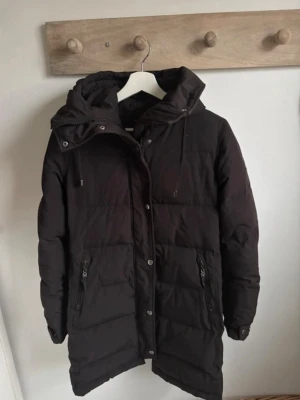 Svart lång dunjacka från Hollies - Säljer en svart lång dunjacka från Hollies med huva och dragkedja framtill. Jackan har två fickor med dragkedja, tryckknappar vid ärmsluten och quiltad design. Perfekt för kalla dagar och riktigt skön att bära.