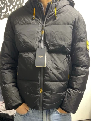 Svart pufferjacka från Fendi - Svart pufferjacka från Fendi med gul logotyp på ärmen och detaljer i gult på dragkedjan och snörningen. Jackan har huva, lång ärm och quiltad design. Materialet är syntet och jackan har en normal passform. Perfekt för kalla dagar.