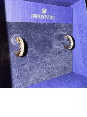 Swarovski guldörhängen med kristaller - Säljer ett par snygga guldpläterade örhängen från Swarovski med gnistrande vita kristaller. Designen är halvcirkelformad och örhängena har en lyxig vibe med tydliga stenar som ger extra bling. Perfekt för dig som gillar statement-smycken och vill sticka ut.