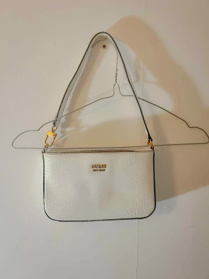 Vit axelväska från Guess med croco-mönster - Säljer en snygg vit axelväska från Guess med croco-mönstrad yta och guldfärgade detaljer. Väskan har dragkedja upptill och en extra ficka med dragkedja på baksidan. Insidan är beige med en innerficka. Perfekt storlek för det viktigaste.