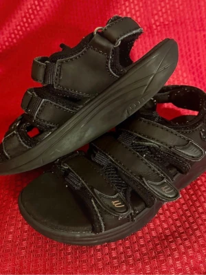 JOBI arbetsandaler  svarta  med kardborreband 38 - Svarta sandaler med flera justerbara kardborreband och robust platt sula. Skorna har detaljer i syntetmaterial och mesh för extra komfort och ventilation. Perfekt för varma dagar och enkel att ta på tack vare smidiga remmar.