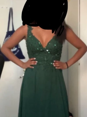 Grön långklänning med spets och paljetter - Säljer en elegant grön långklänning med djup v-ringning och öppen rygg. Toppen har dekorativa paljetter och spetsdetaljer, medan kjolen är flödande och enfärgad. Klänningen är ärmlös och har meshpartier vid axlar och rygg för extra detalj.