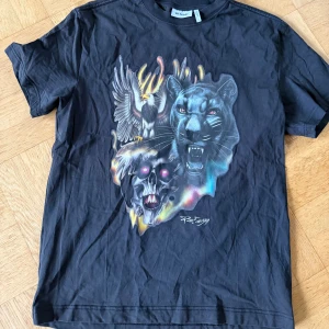 Svart t-shirt från Weekday - Cool svart t-shirt från Weekday med ett fett tryck på framsidan som visar en panter, en örn och en dödskalle med neonfärger. T-shirten har rund hals och korta ärmar, gjord i mjuk bomull som är skön mot huden.