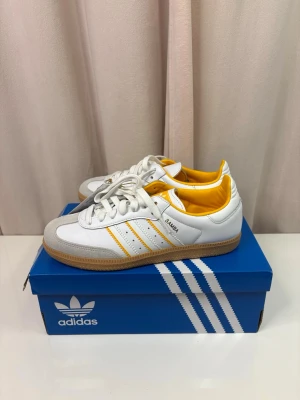 Adidas Samba sneakers vit/gul - Adidas Samba sneakers i vitt skinn med gula detaljer och klassiska tre ränder på sidan. Yttersulan är i gummi och tån har en grå mockadetalj. Insidan är fodrad i gul färg och skorna har snörning. Perfekt för dig som gillar retrostil och sportig vibe.