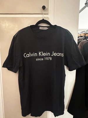 Svart t-shirt Calvin Klein Jeans M - Svart t-shirt från Calvin Klein Jeans med vit text 'Calvin Klein Jeans since 1978' på bröstet. Klassisk rund hals och korta ärmar. Regular fit för en skön och avslappnad passform. Materialet är mjuk bomull som känns nice mot huden.