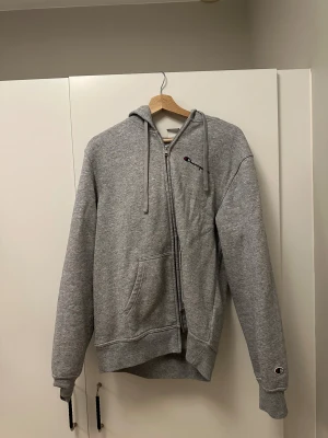 En grå Champion kofta - En grå champion kofta med luva. Dragkedjan är lite trasig då den inte går ner hela vägen (sista bilden visar). Det går möjligtvis att fixa dragkedjan.  Utöver detta är koftan i bra skick. 
