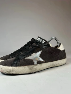 Svarta GGDB Superstar sneakers mocka - Säljer ett par GGDB Superstar sneakers i svart mocka med vita detaljer och silverfärgad stjärna på sidan. Skorna har svarta skosnören, vit hälkappa och GGDB-logga på plösen. Klassisk låg modell med slitna detaljer och platt sula.