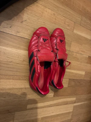 Röda Adidas fotbollsskor med svarta ränder - Säljer ett par röda Adidas fotbollsskor med klassiska svarta ränder på sidan. Skorna har snörning och en täckande plös med Adidas-logga. Tillverkade i skinn för bra känsla och komfort på planen. Perfekta för dig som vill sticka ut med färg och stil under matchen.