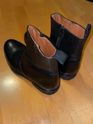 Svarta läderkängor med dragkedja - Svarta boots i äkta skinn med grov sula och dragkedja på sidan. De är tillverkade av äkta läder i Spanien för hand. Helt nya och oanvända men saknas låda och lappar. Insidan är fodrad i brunt läder och yttersulan har tydligt mönster för extra grepp. Klassisk rund tå och praktisk flik bak för enkel påtagning. Nypris 1975kr så säljs billigt. Priset är inte hugget i sten. Skriv vid minsta fråga! 