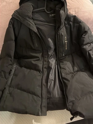 Svart pufferjacka från Sail Racing  - Svart pufferjacka från Sail Racing med huva och flera praktiska fickor med dragkedja. Jackan har en stilren design och logotyp på bröstet samt ärmen. Perfekt för kalla dagar och har en skön, oversized passform.