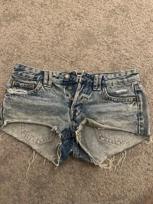 Blå jeansshorts från Pull&Bear - Säljer ett par blå jeansshorts från Pull&Bear i storlek 36. De är högmidjade med fransiga kanter och slitna detaljer, klassisk femficksmodell och knappar framtill. Perfekta för varma dagar och har en cool vintagekänsla.