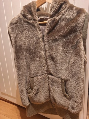 Brun pälsväst med huva och fickor - Säljer en brun pälsväst med huva och två stora fickor framtill. Västen har dragkedja och ribbstickade detaljer vid fickor och nederkant. Perfekt för lager-på-lager och extra mysig tack vare det fluffiga materialet.