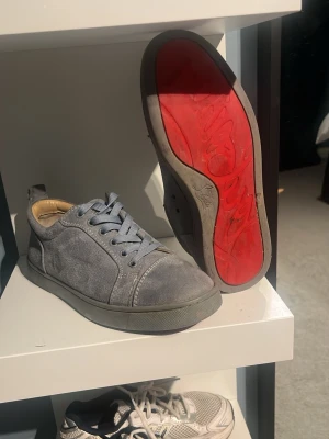 Grå mockasneakers med röd sula - Säljer ett par grå sneakers i mocka från Christian Louboutin med ikonisk röd sula. Skorna har rund tå, snörning i matchande grått och diskret söm längs kanterna. Insidan är beige och sulan är platt för en clean look.
